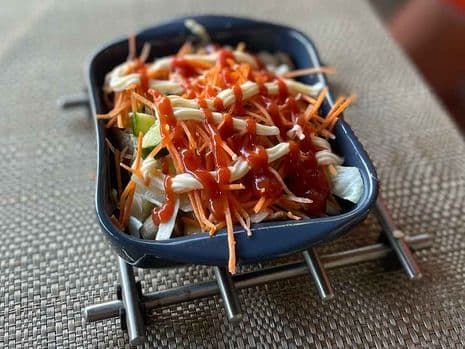Een kapsalon met gesneden wortelen, kool en saus bovenop, geserveerd in een rechthoekig zwart dienblad op een geweven placemat.