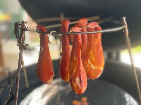 Verschillende stukken gerookte kipfilet hangend aan haken in een kamado barbecue, met een onscherpe achtergrond.