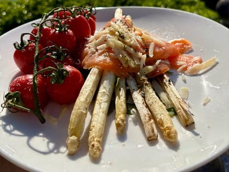 Witte asperges belegd met gerookte zalm en parmezaanse kaas, vergezeld met trostomaatjes, geserveerd op een wit bord.