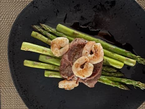 Surf en turf van gegrilde ossenhaas met garnalen op een bedje van asperges, geserveerd op een zwart bord.