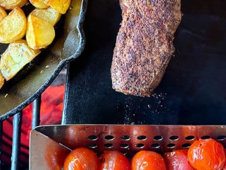 Diamanthaas wordt gegrild op een speksteen, omringd door gebakken aardappelen in een koekenpan en geroosterde tomaten in een metalen mandje.