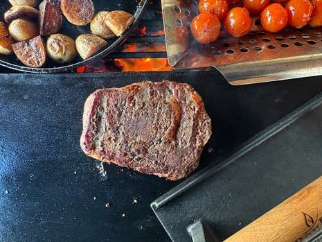 Een gegrilde ribeye op een speksteen, met daarnaast geroosterde aardappelen en cherrytomaatjes