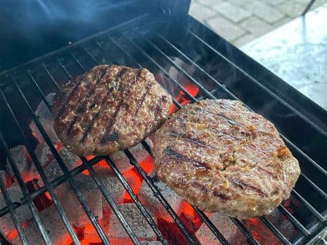 Twee runderburgers met grillmarkeringen bakken boven gloeiend hete houtskool op een barbecuerooster, met opstijgende rook.