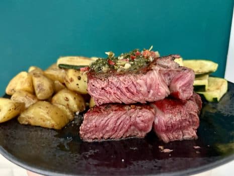 Close-up van een bord met medium-rare ribeye met daarop chimichurri, geroosterde aardappelen en gegrilde courgette.