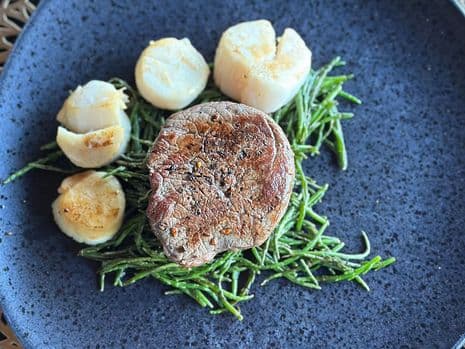 Een geschroeide tournedos en coquilles op een bedje van zeekraal, geserveerd op een zwart bord.