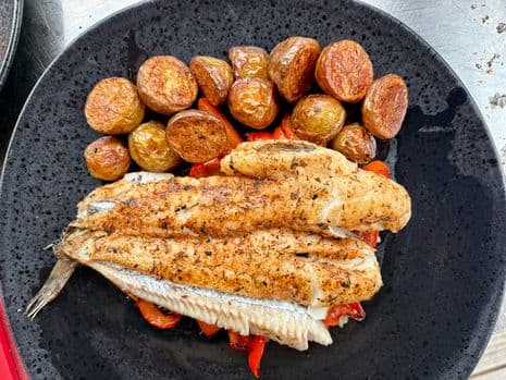 Gegrilde wijting op een zwart bord met geroosterde krieltjes en rode paprika.