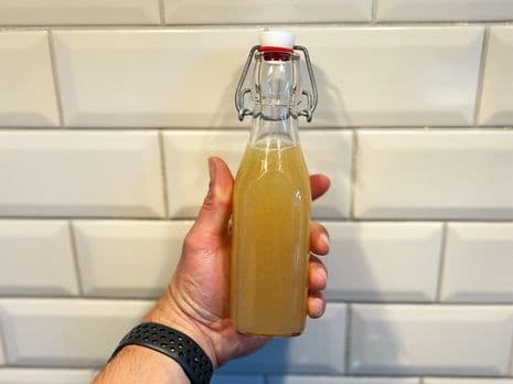 Hand met een glazen fles gevuld met grilled lemonade tegen een witte betegelde achtergrond.