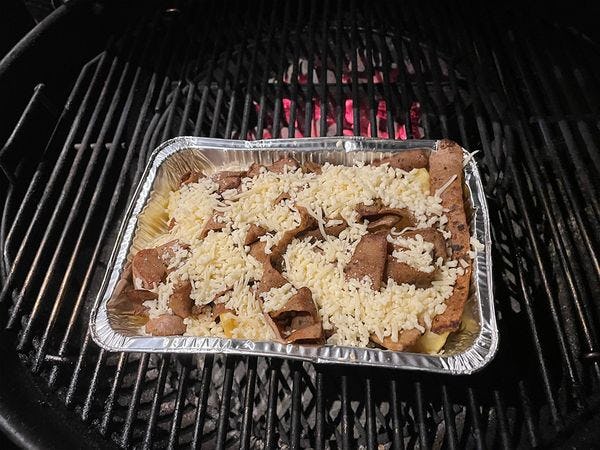Een aluminiumbakje met daarin friet en opgebakken frikandellen linten met kaas op een barbecuerooster