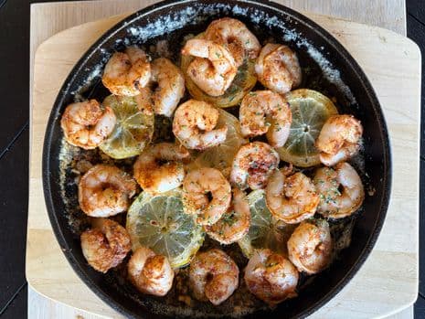 Een skillet met gekruide garnalen en schijfjes citroen, gegarneerd met kruiden en knoflookboter op een houten plank.