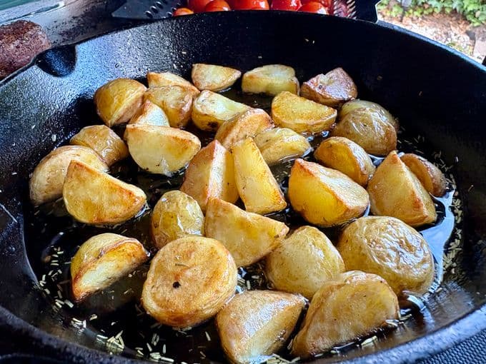 Goudbruin geroosterde aardappelen in een skillet, glanzend van de olie en kruiden, met tomaten op de achtergrond.